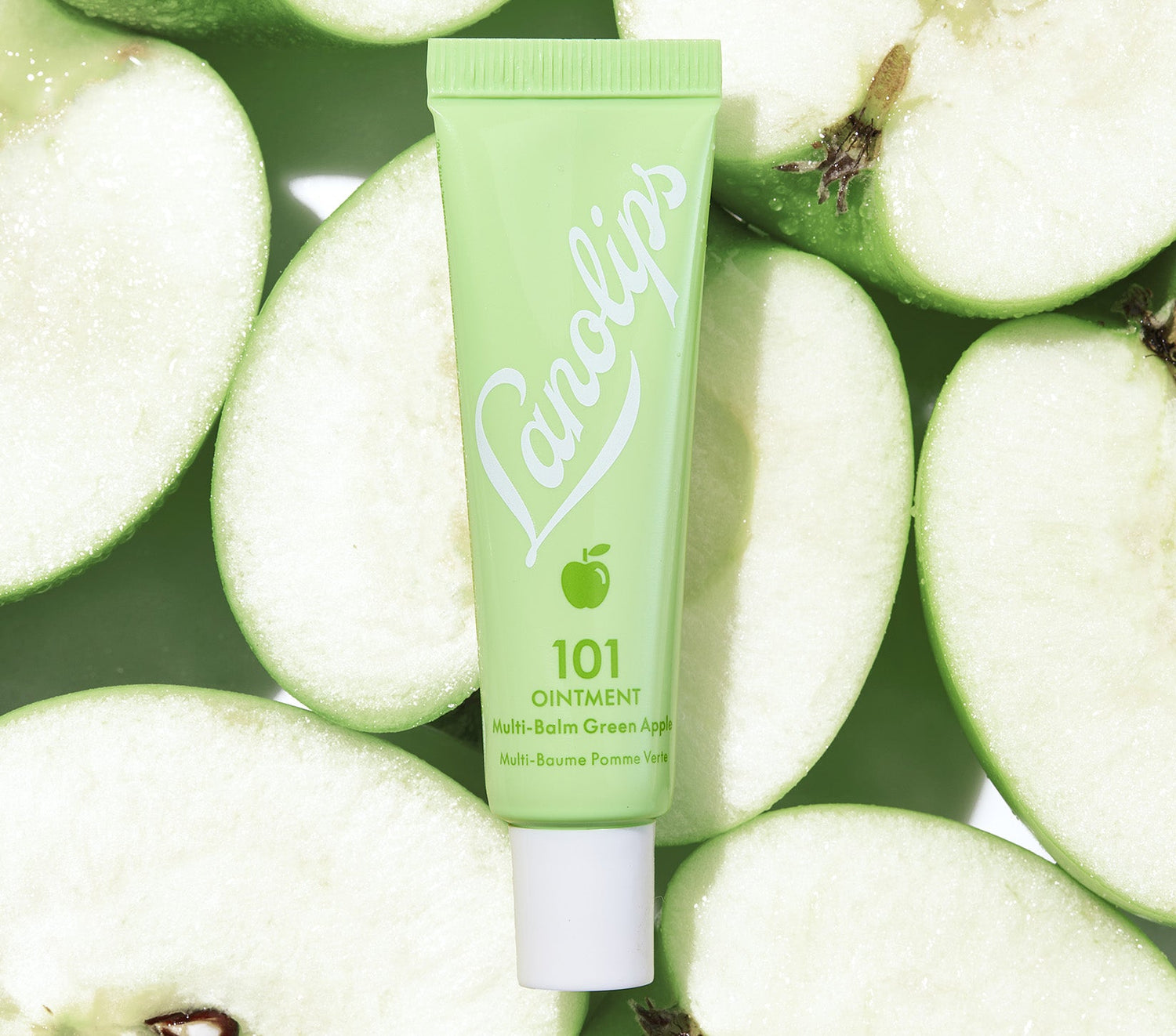 A tube of Lanolips 101 Ointment in the limited-edition Green Apple flavour is placed on a background of freshly sliced Granny Smith apples. The tube is a soft green shade with white branding and a small apple icon, reflecting the crisp and refreshing theme of the product. This nourishing ointment contains Aussie lanolin, vitamin e and green apple extract, which mimics the skin’s natural oils to provide intense hydration, leave lips feeling nourished.