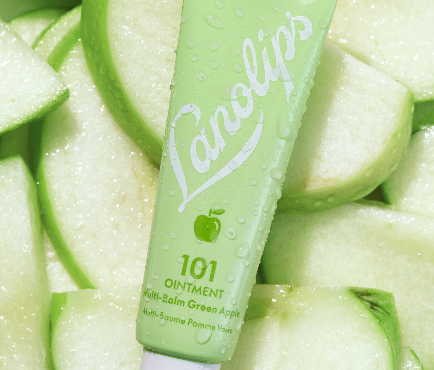 A tube of Lanolips 101 Ointment Multi-Balm in Green Apple rests on a bed of fresh Granny Smith apples. The tube is a vibrant green shade, feature the white Lanolips logo and a small apple icon, complimenting the crispy, juicy apples around it. The product contains lanolin and vitamin-e, deeply hydrating ingredients that mimic the skin's natural oils, as well as provide long lasting hydration on dry lips.