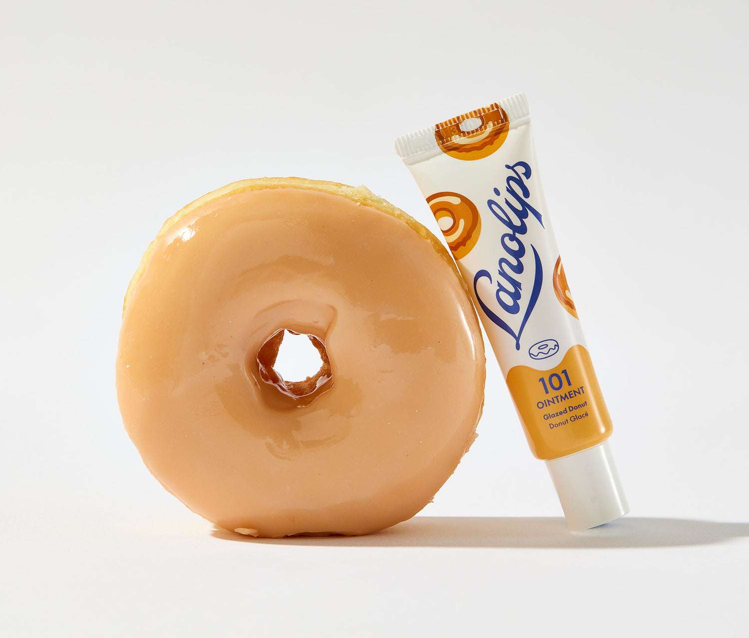 A tube of Lanolips 101 Ointment Multi Balm in Glazed Donut leaning against a donut. The tube features a soft yellow and white design, with blue Lanolips logo branding and illustrations of donuts. The product contains Aussie lanolin, a deeply hydrating and nourishing ingredients that mimics the skin's natural oils. It also locks in moisture and provides a glossy, long lasting finish.