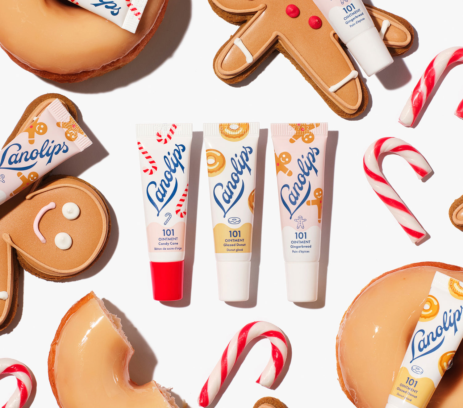 Lanolips 101 Ointment Holiday Mini Bites lip balm trio in Candy Cane, Gingerbread and Glazed Donut flavours, packed with rich lanolin, vitamin E and natural flavors, styled with gingerbread cookies, donuts and candy canes for a holiday twist.
