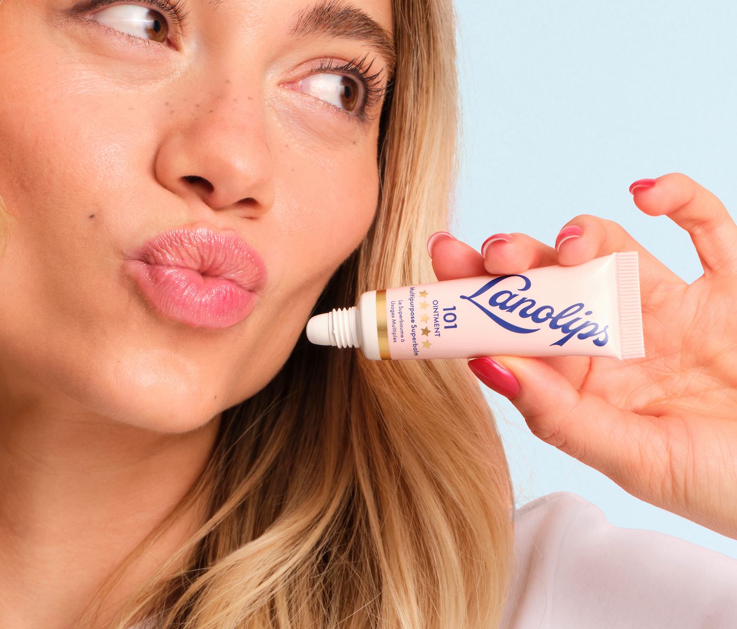Model holding a Lanolips Original 101 Ointment Multipurpose Superbalm, with a blue background. Cult-classic, dermatologically tested balm for lips, skin, elbows and cuticles.