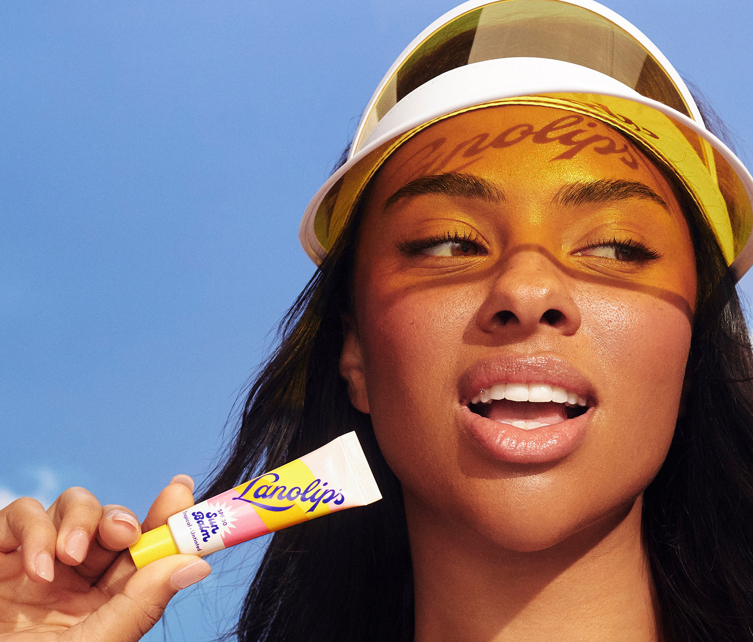 A model holds a tube of Lanolips Sun Balm SPF 30 Tropical lip balm against a blue sky backdrop. Wearing a white-and-yellow sun visor, the word “Lanolips” is cast as a warm shadow across her forehead. Her glossy lips and radiant skin reflect the balm’s protective, hydrating, and summery feel - perfectly capturing SPF-meets-skincare for everyday glow.