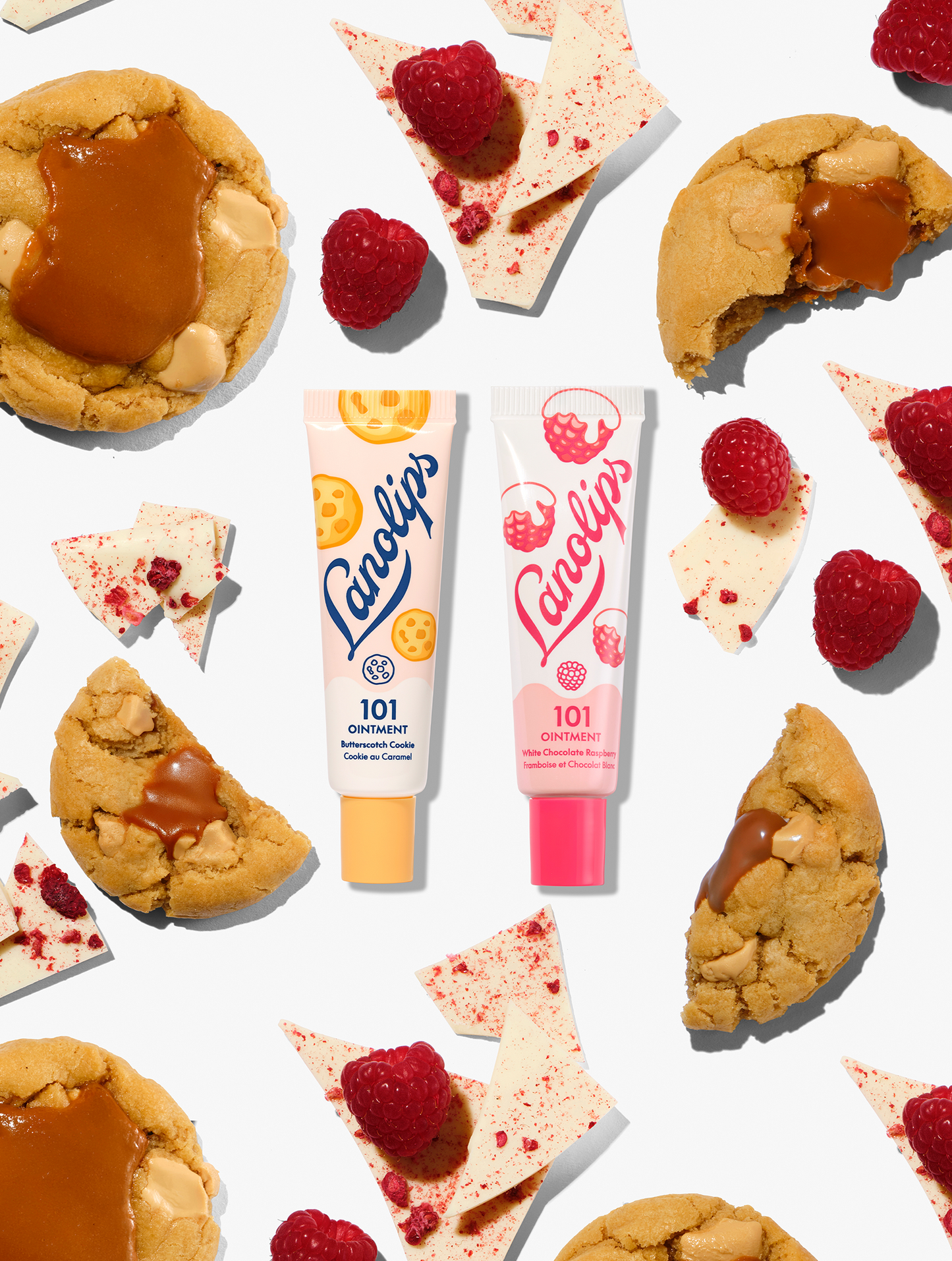 Lanolips 101 Delicious collection duo featuring 101 Ointment Multi-Balms in Butterscotch Cookie and White Chocolate Raspberry. The two natural lanolin balm tubes are pictured together amongst chewy caramel biscuits, fresh raspberries, and shards of white chocolate.