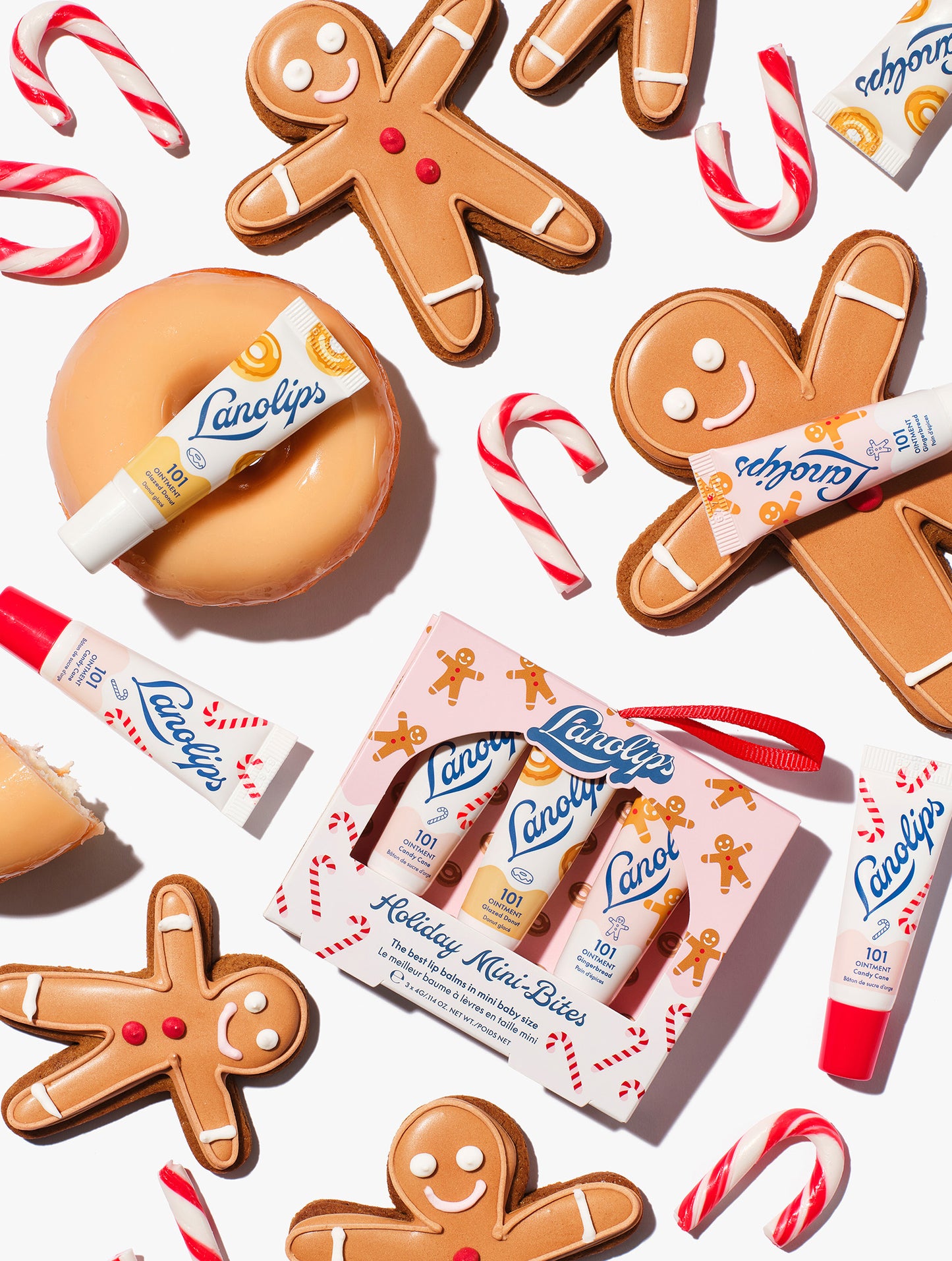 Flatlay of Lanolips Holiday Mini Bites lip balm trio gift set with Candy Cane, Gingerbread and Glazed Donut flavors, styled with gingerbread cookies and candy canes.