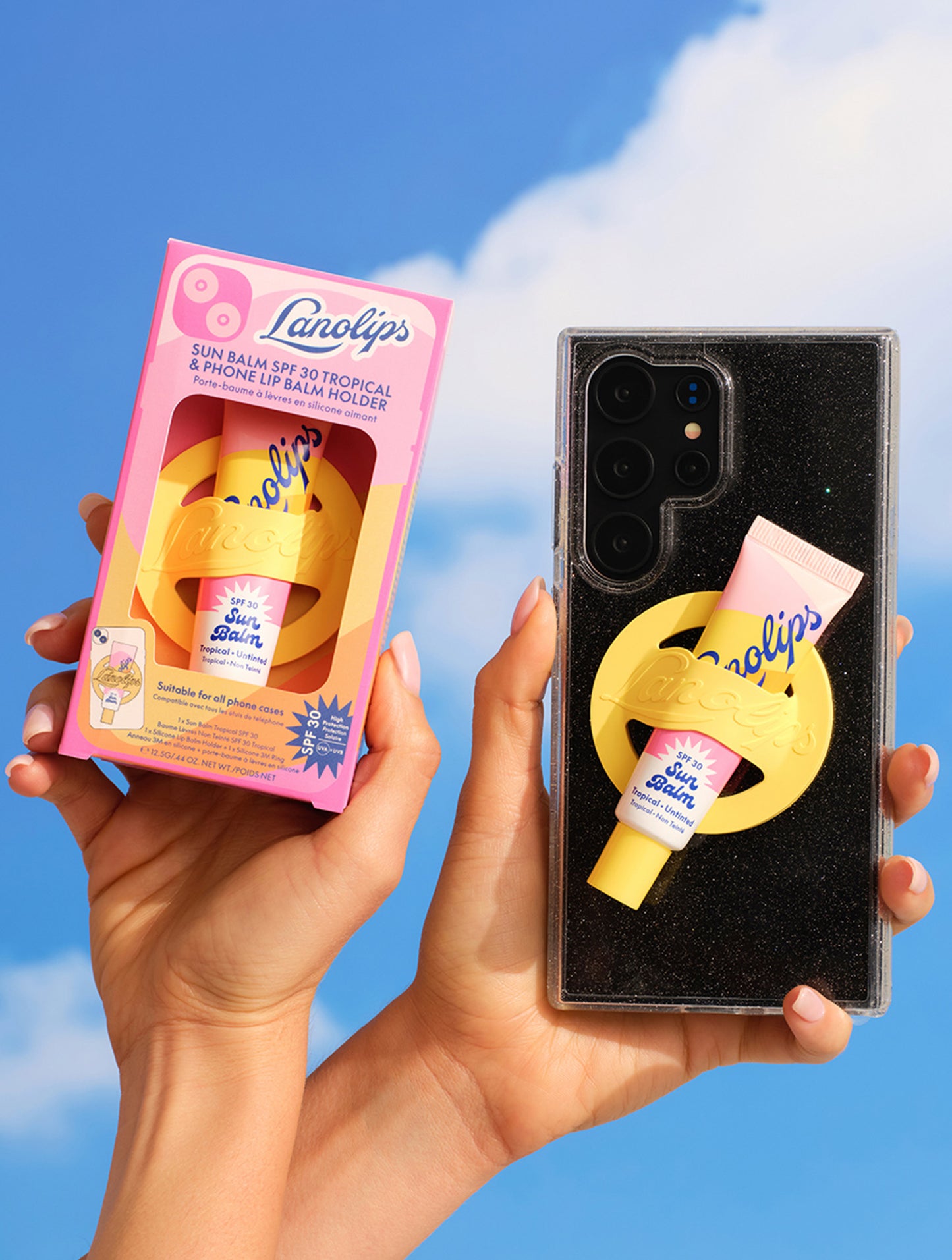 Lanolips Sun Balm SPF 30 Tropical with yellow phone holder shown on a clear glitter phone case, alongside its pink and yellow packaging, held up against a bright blue sky.