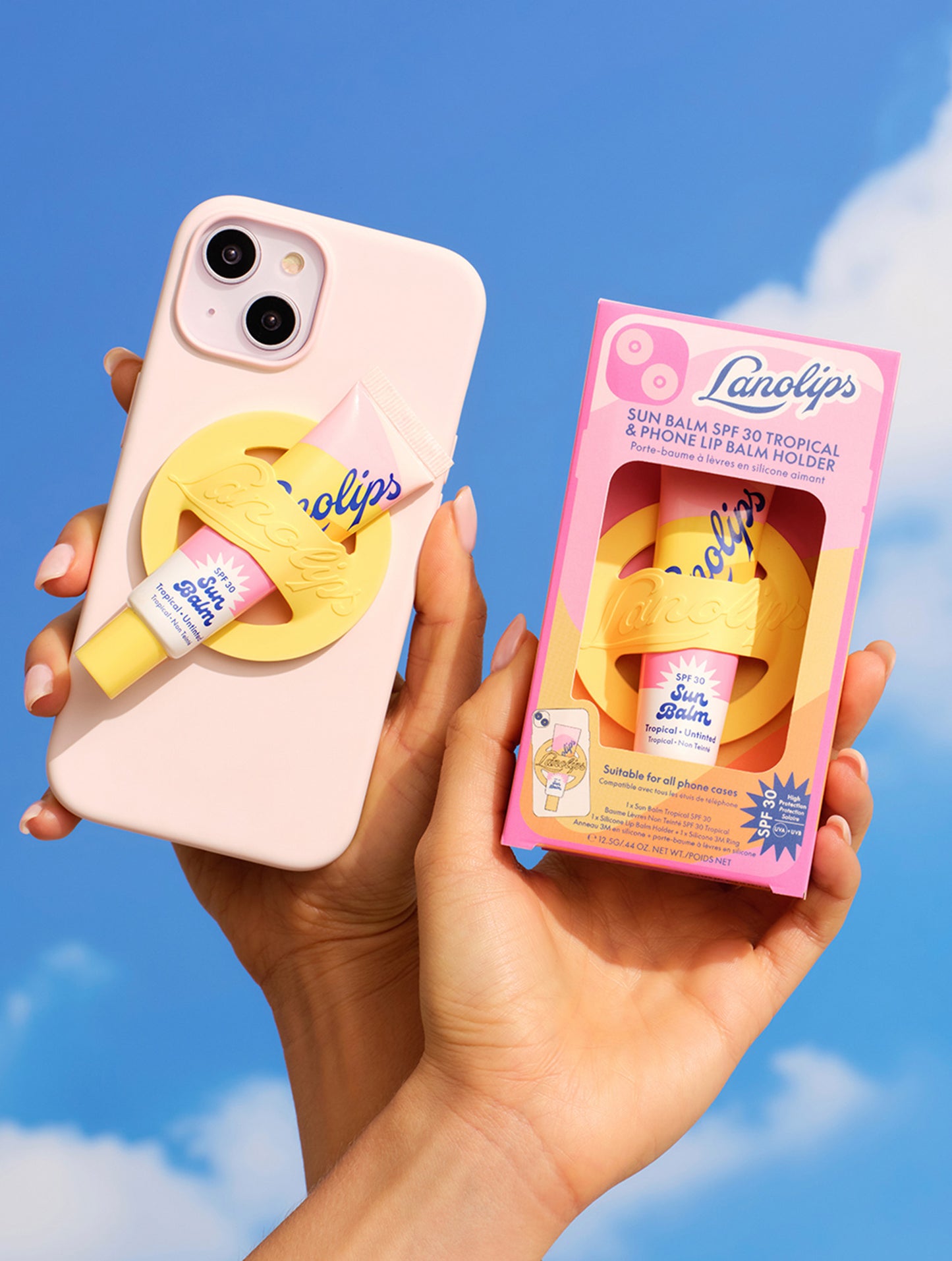 A hand holds a pastel pink phone with a yellow Lanolips Sun Balm SPF 30 phone lip balm holder securely attached, featuring a pink tube of Lanolips Sun Balm SPF 30 Tropical. Beside it, another hand displays the matching boxed set against a sunny blue sky. A fun and functional phone accessory for lip balm on-the-go.