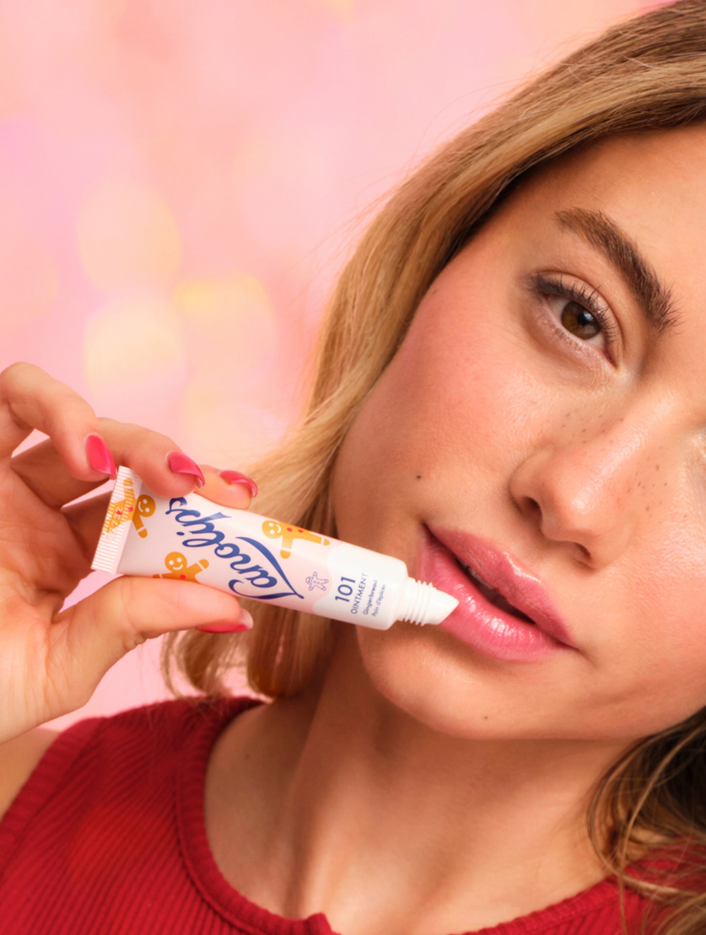 Model applying Lanolips 101 Ointment Multi-Balm Gingerbread lip balm to her lips with a pink background. Embrace warm spice and soft lips with this limited edition festive 101 Delicious Holiday gourmand flavor.