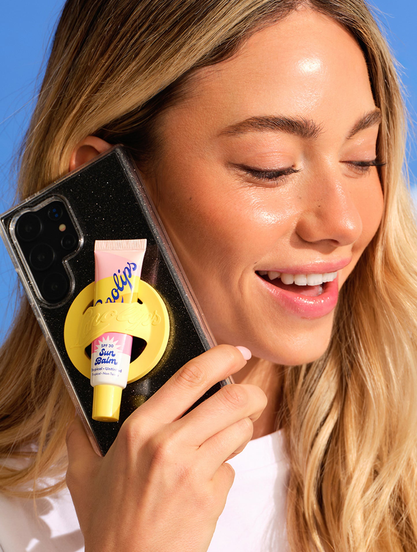 Model holding a phone with a tube of Lanolips Sun Balm SPF 30 Tropical Lip Balm and yellow lip balm holder against a blue sky