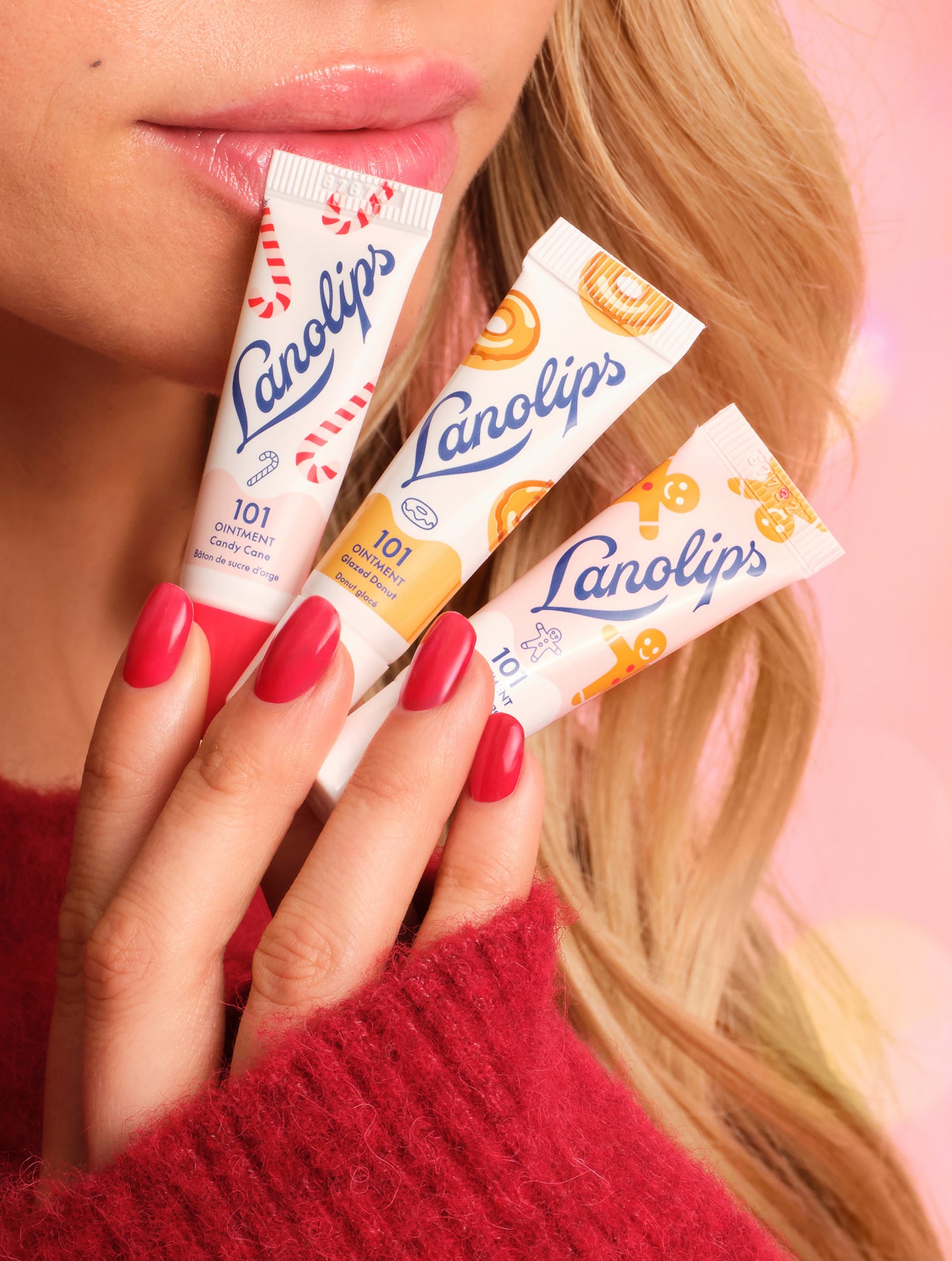 Close-up of model holding Lanolips Holiday Mini Bites lip balm trio in Candy Cane, Gingerbread and Glazed Donut flavors, with glossy lips and red nails.