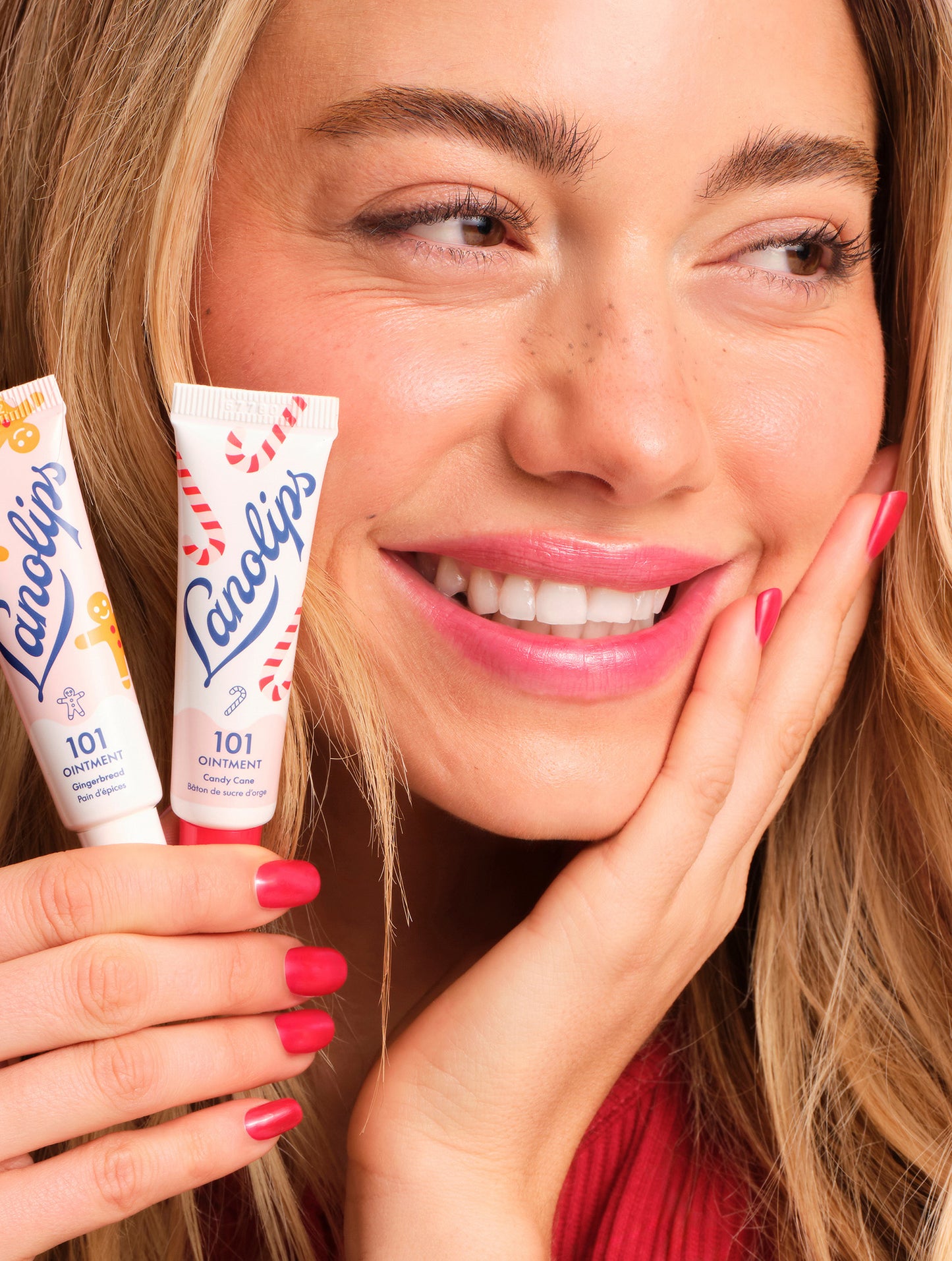 Model smiling while holding Lanolips 101 Ointment Multi-Balm Gingerbread & Candy Cane, made with lanolin, vitamin E and natural flavors, from the limited-edition Holiday Gingerbread House collection.