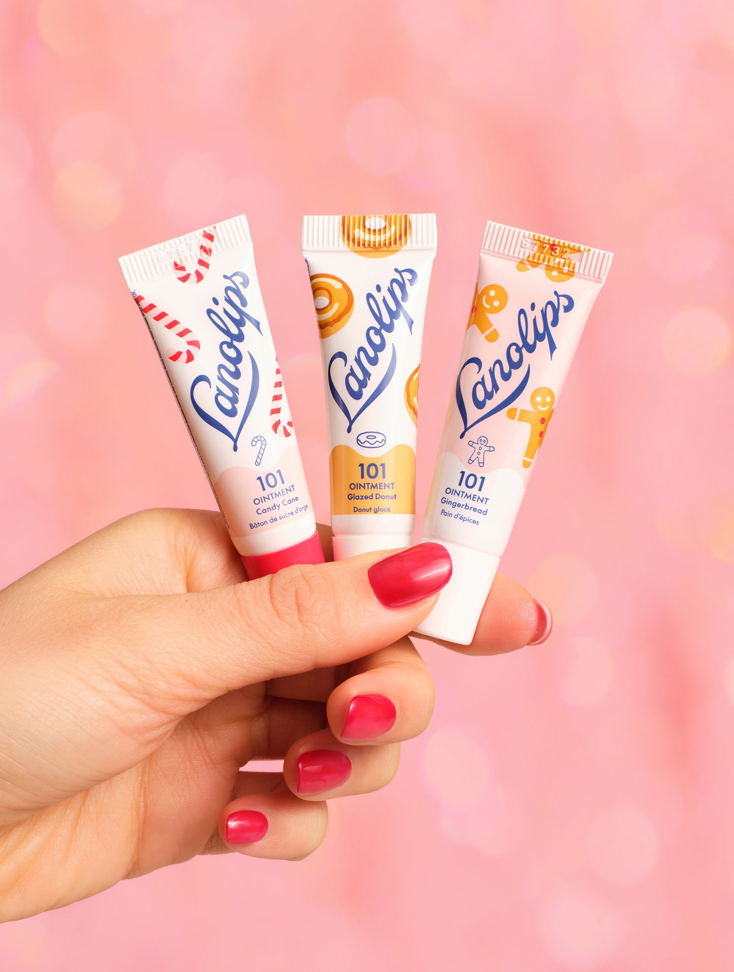 Model holding the trio from the Lanolips Holiday Mini Bites set. The set contains the two new festive flavors - gingerbread and candy cane, as well as a fan-favorite, glazed donut. Packed with rich lanolin, vitamin E, and gourmand flavor, this holiday trio delivers deep hydration for lips, cuticles, dry patches & more.