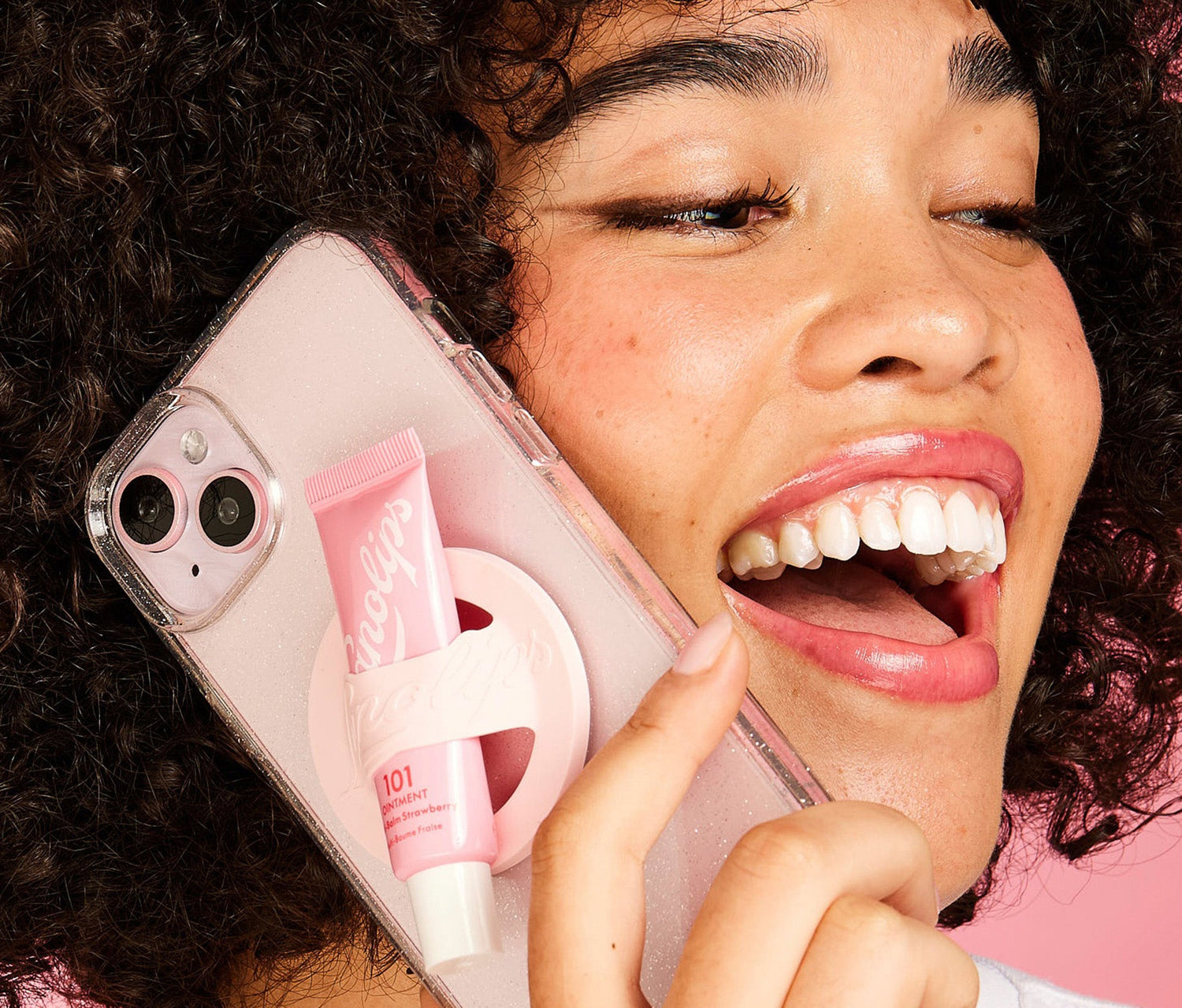 A model holding a pink phone to their ear. Attached to the back of the phone case is a small tube of the Lanolips 101 Ointment Multi Balm in Strawberry, secured with the Lanolips pink phone lip balm holder.