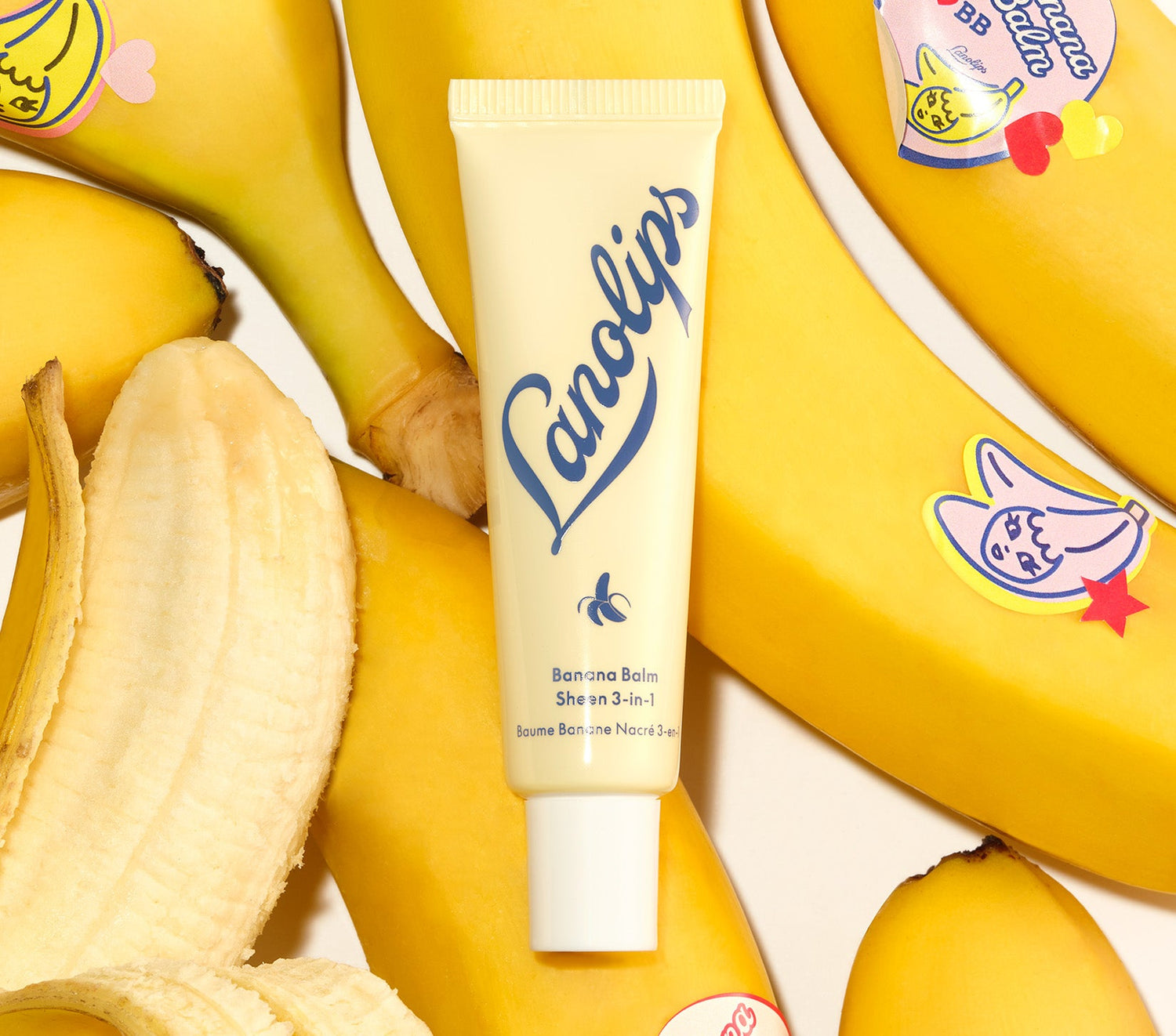 A tube of Lanolips Banana Balm Sheen 3-in-1, in soft yellow packaging with blue lettering and a small banana icon, placed among fresh bananas. Some bananas have playful stickers, while one is partially peeled, revealing its creamy interior. Made with lanolin, cocoa seed butter, banana extract, and a hint of shimmer, this balm provides deep hydration for glossy lips.