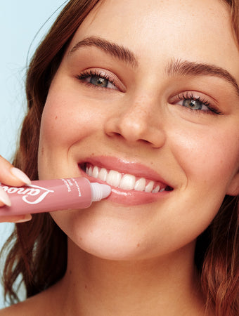 A smiling young woman with dewy skin and wavy light brown hair applies Lanolips Tinted Lip Balm in Perfect Nude to her full lips. The hydrating lip balm gives a glossy, natural pink tint, enhancing her radiant complexion.The close-up shot captures the balm’s nourishing texture and smooth application, making it the perfect beauty essential for soft, hydrated lips.