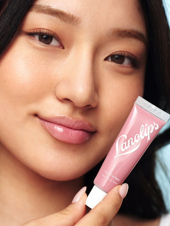 A close-up of a woman with smooth, radiant skin and long, dark hair holding Lanolips Tinted Lip Balm in Rhubarb near her face. Her full lips are coated in the nourishing balm, delivering a sheer, rosy pink tint with a glossy, hydrating finish. The pastel pink packaging of the Lanolips tube, featuring white branding, complements the soft yet vibrant lip color.  This ultra-hydrating tinted lip balm enhances lips with a healthy flush while keeping them soft and moisturized. 