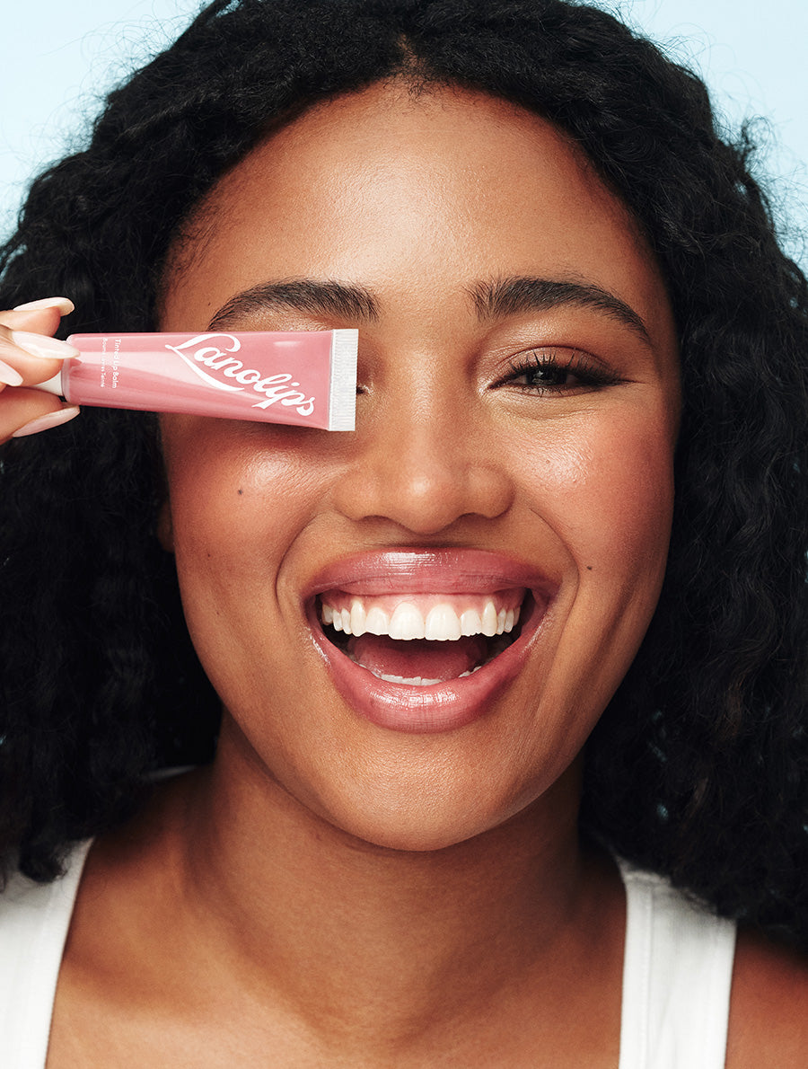 Model with glowing deep-toned skin and voluminous curls holds a tube of Lanolips Tinted Lip Balm in Rhubarb playfully over one eye while smiling wide. Her lips are coated in the nourishing, sheer rosy-pink balm, giving them a glossy, plump, and hydrated finish. The pastel blue background enhances the vibrant yet effortless beauty look. This ultra-hydrating tinted lip balm delivers a dewy shine and a healthy, moisturized feel.