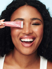 Rose Tinted Lanolin Lip Balm | Load image into Gallery viewer, Model with glowing deep-toned skin and voluminous curls holds a tube of Lanolips Tinted Lip Balm in Rhubarb playfully over one eye while smiling wide. Her lips are coated in the nourishing, sheer rosy-pink balm, giving them a glossy, plump, and hydrated finish. The pastel blue background enhances the vibrant yet effortless beauty look. This ultra-hydrating tinted lip balm delivers a dewy shine and a healthy, moisturized feel.

