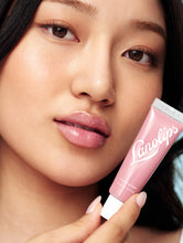 Rose Tinted Lanolin Lip Balm | Load image into Gallery viewer, Model with glowing deep-toned skin and voluminous curls holds a tube of Lanolips Tinted Lip Balm in Rhubarb playfully over one eye while smiling wide. Her lips are coated in the nourishing, sheer rosy-pink balm, giving them a glossy, plump, and hydrated finish. The pastel blue background enhances the vibrant yet effortless beauty look. This ultra-hydrating tinted lip balm delivers a dewy shine and a healthy, moisturized feel.
