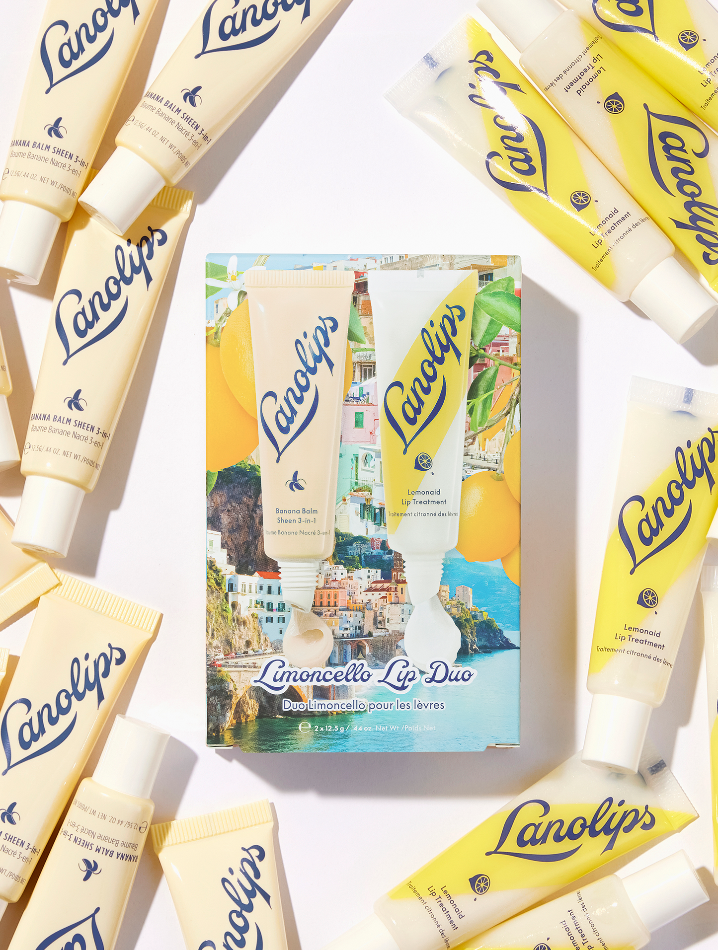 Lanolips Limoncello Lip Duo set features a tube of banana balm lip sheen and a tube of lemonaid lip treatment against a scenic Mediterranean coastal collage background with lemons.