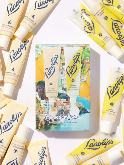 Lanolips Limoncello Lip Duo set features a tube of banana balm lip sheen and a tube of lemonaid lip treatment against a scenic Mediterranean coastal collage background with lemons.