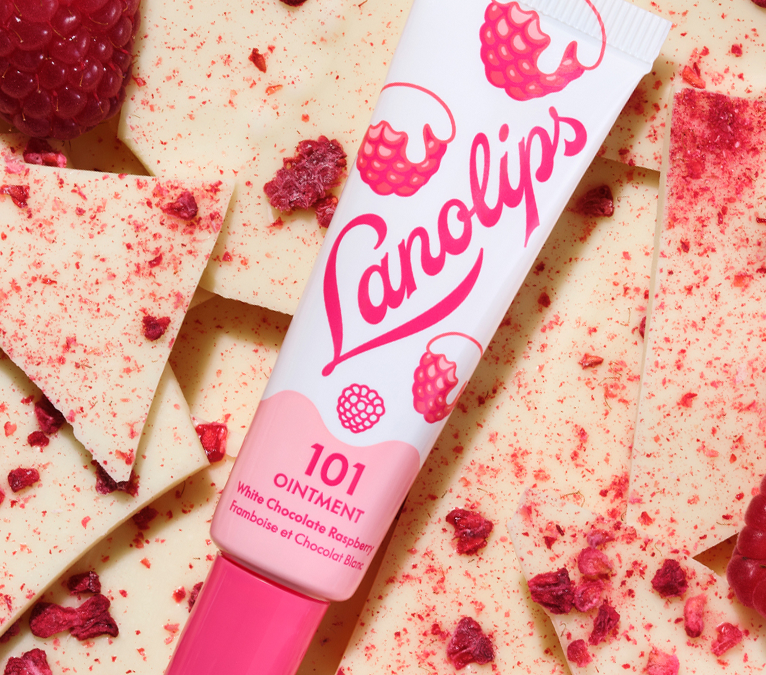 A close-up of Lanolips 101 Ointment White Chocolate Raspberry resting on creamy white chocolate shards and dried raspberry pieces. This 100% natural lanolin multi-balm is infused with Vitamin E and all-natural flavours to provide deep, long-lasting hydration.