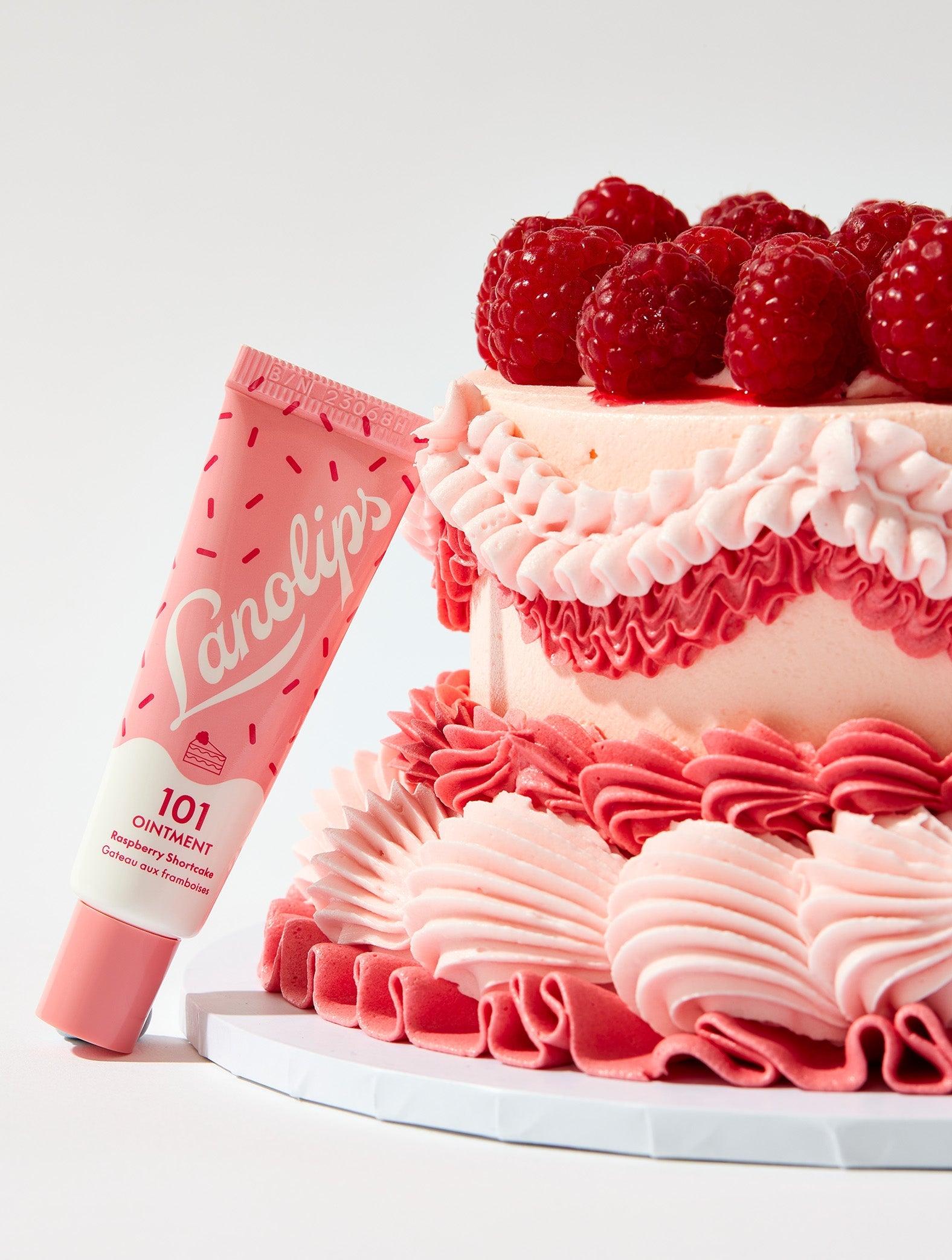 Lanolips 101 Ointment Multi-Balm in Raspberry Shortcake flavour, leaning next to a pink vintage cake topped with fresh raspberries. Made with ultra-rich lanolin, all-natural raspberry flavour and vitamin e, this balm delivers intense hydration for lips, cuticles and dry skin.