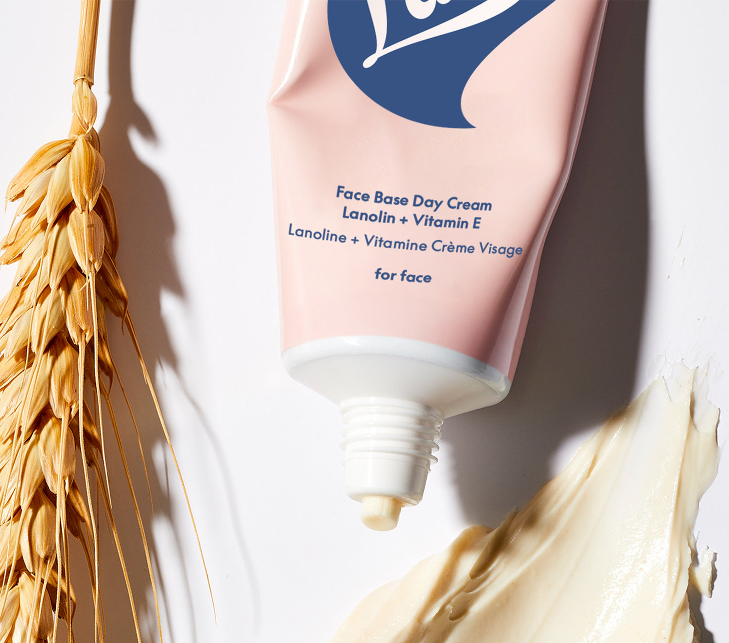 Close up shot of a tube of Lanolips Face Base Day Cream Lanolin + Vitamin E with wheat stalk and cream smear on a light background.
