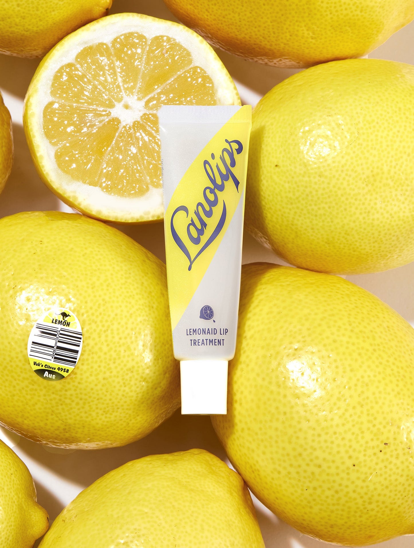 Lanolips Lemonaid Lip Treatment in a sleek, squeezable tube with a white cap and a bright yellow diagonal design. The packaging features the Lanolips logo in bold blue lettering and an illustration of a lemon slice, highlighting its citrus-infused, ultra-hydrating formula. The lip treatment has a glossy, rich texture designed to nourish, exfoliate, and soften dry lips with a refreshing lemon scent.