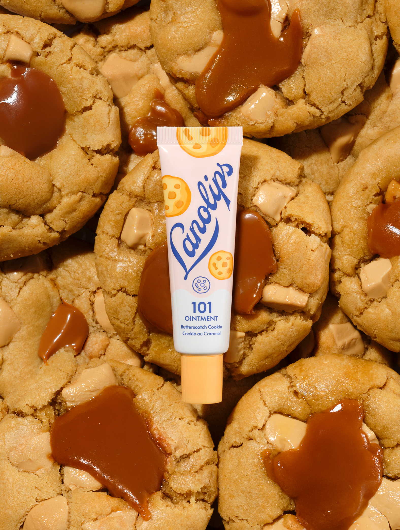 Lanolips 101 Multi-Balm Ointment in Butterscotch Cookie flavour. A 100% natural lanolin balm delivering warm cookie comfort with notes of buttery, golden butterscotch. The tube is pictured atop freshly baked biscuits with gooey caramel, offering deep, long-lasting hydration.