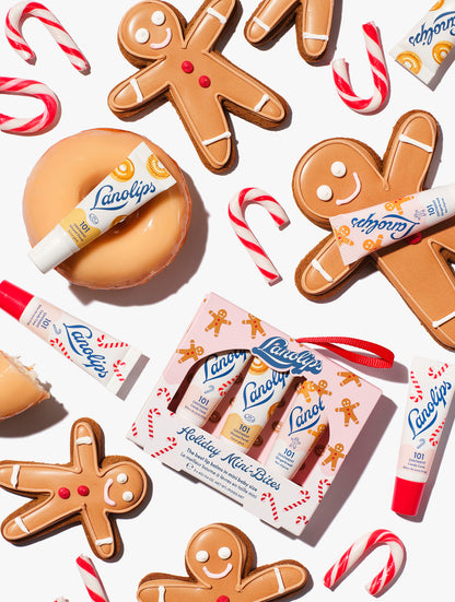 Flatlay of Lanolips Holiday Mini Bites lip balm trio gift set with Candy Cane, Gingerbread and Glazed Donut flavors, styled with gingerbread cookies and candy canes.