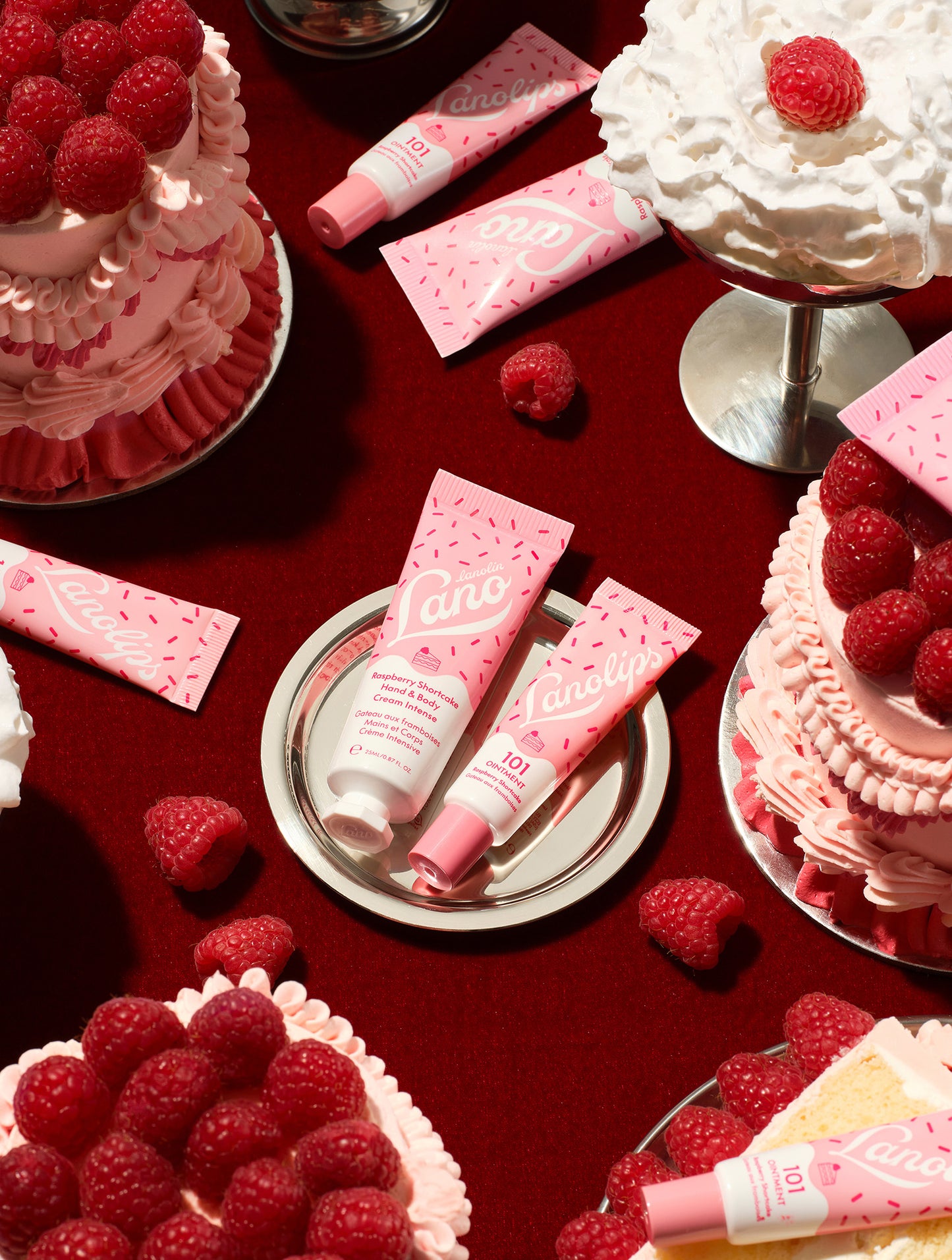 Lanolips Raspberry Shortcake Holiday Duo – 101 Ointment Multi-Balm and Hand & Body Cream Intense, styled with raspberry shortcake, whipped cream and fresh raspberries on a red background.