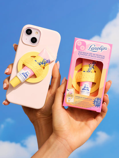 A hand holds a pastel pink phone with a yellow Lanolips Sun Balm SPF 30 phone lip balm holder securely attached, featuring a pink tube of Lanolips Sun Balm SPF 30 Tropical. Beside it, another hand displays the matching boxed set against a sunny blue sky. A fun and functional phone accessory for lip balm on-the-go.