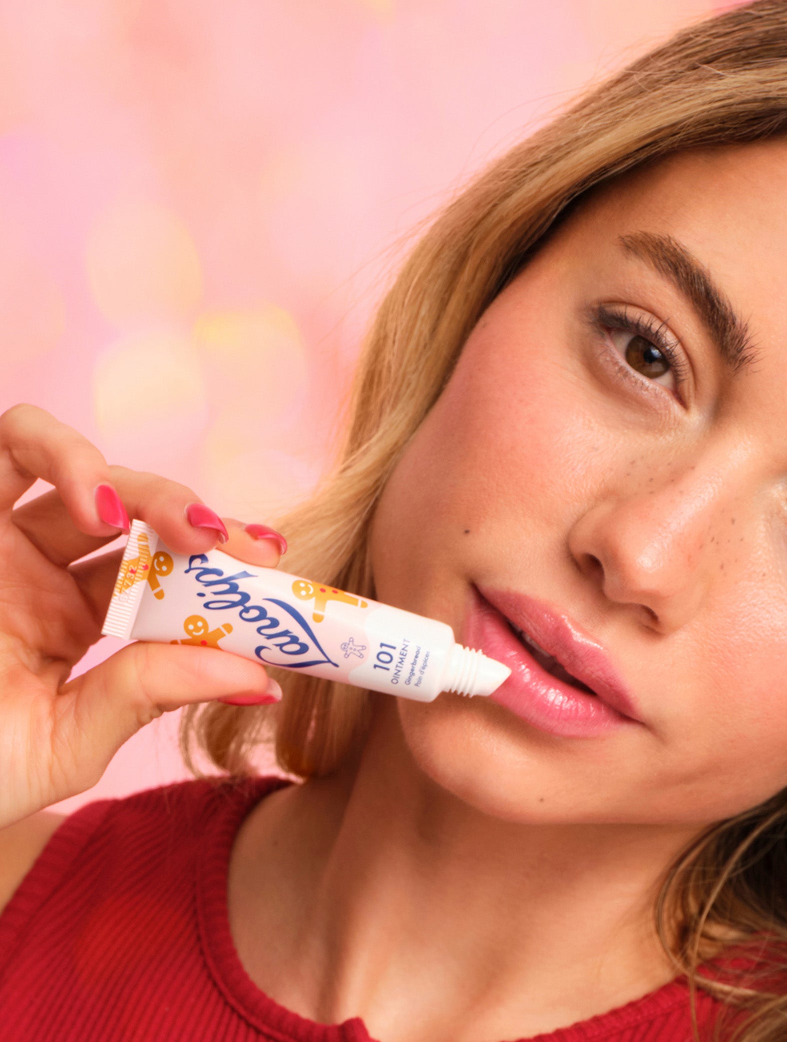 Model applying Lanolips 101 Ointment Multi-Balm Gingerbread lip balm to her lips with a pink background. Embrace warm spice and soft lips with this limited edition festive 101 Delicious Holiday gourmand flavor. 