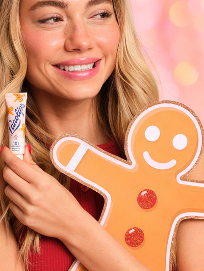 Model holding a cut out of the gingerbread man cut out and a Lanolips 101 Ointment Multi-Balm Gingerbread against a colorful background.
